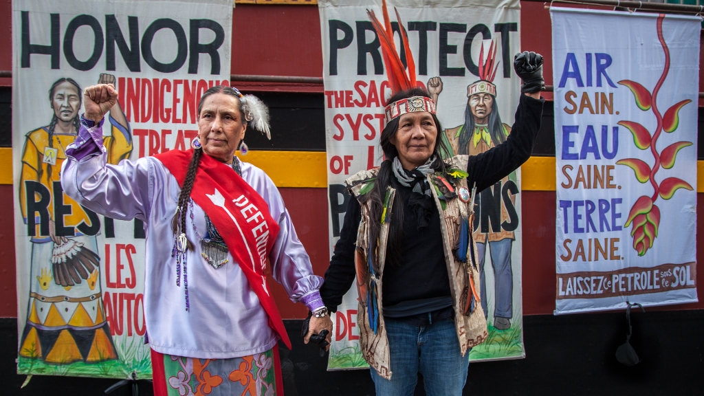Casey Camp Horinek (Oklahoma) and Gloria Ushigua Santi (Sapara, Ecuador) standing in mutual solidarity (in front of their respective portraits by Mona Caron)